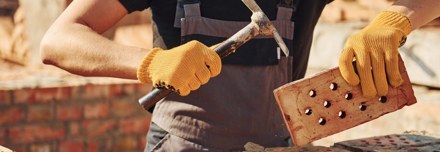 Holding brick and using hammer. Construction worker in uniform and safety equipment have job on building.