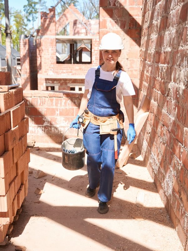 Hardworking woman building engineer in uniform and toolbelt carrying bucket of mortar for laying bricks on construction site, building houses