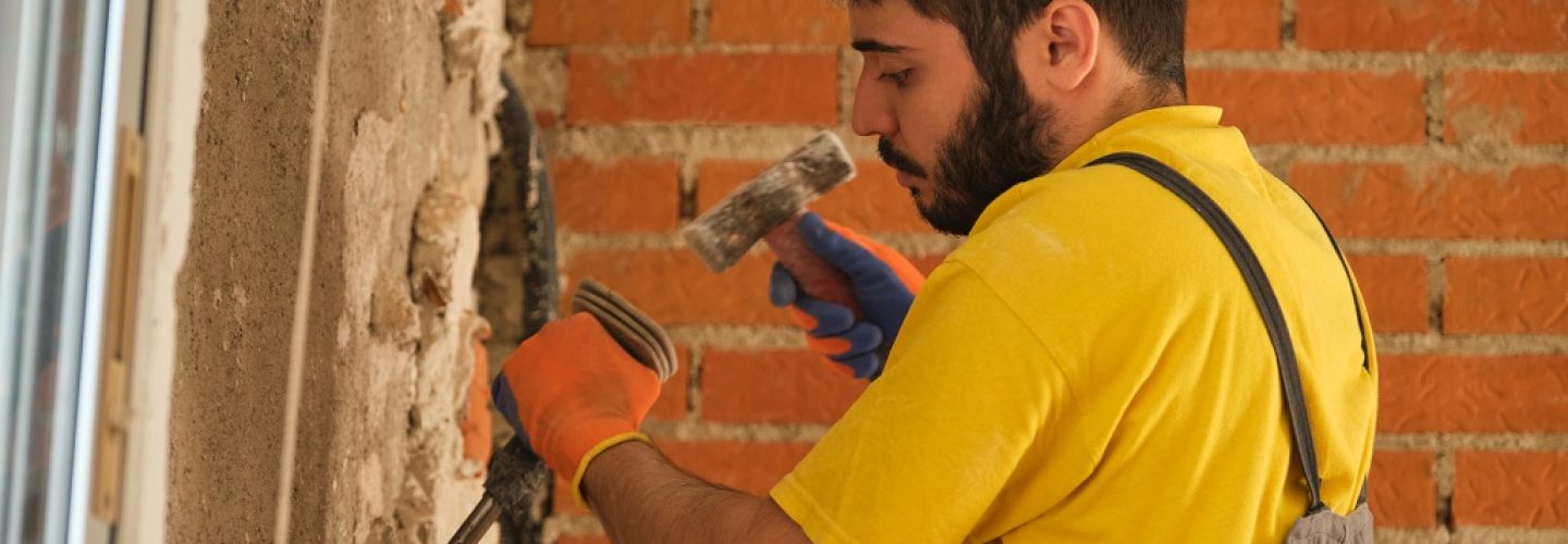 Young caucasian construction worker breaking up a house wall with a hammer and a chisel.